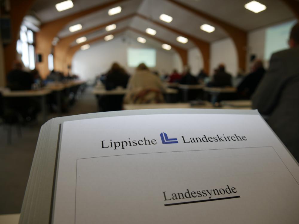 Zukunftssynode - Livestream - Die Lippische Landessynode tagt in Barntrup und Lemgo Im Vordergrund ist eine Mappe zu sehen. Darauf steht: Lippische Landeskirche und dadrunter Landessynode. Im Hintergrund sieht man verschwommen Personen, die in einem großen Raum sitzen.
